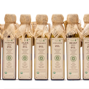 May include: A row of 12 glass bottles with burlap tops, each containing a different type of organic seed oil. The bottles are labeled with the type of oil, including almond, walnut, pumpkin, flax, hazelnut, hemp, mustard, poppy, and black sesame.