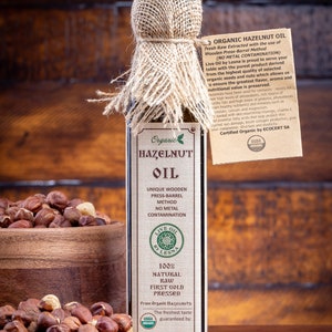 May include: A glass bottle of organic hazelnut oil with a burlap-wrapped cap and a paper tag. The label reads Organic Hazelnut Oil, 100% Natural Raw First Gold Pressed, 8.45 oz (250 ml). A wooden bowl filled with whole hazelnuts sits beside the bottle.