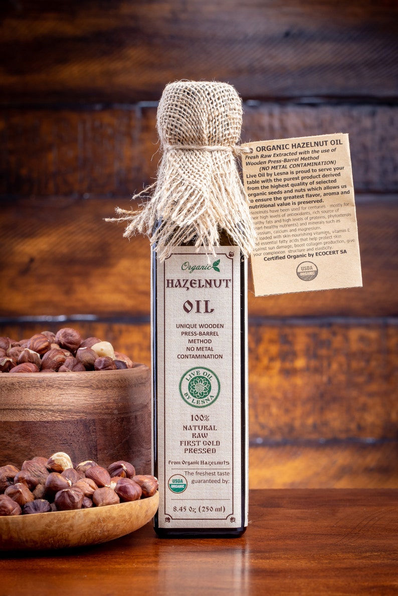 May include: A glass bottle of organic hazelnut oil with a burlap-wrapped cap and a paper tag. The label reads Organic Hazelnut Oil, 100% Natural Raw First Gold Pressed, 8.45 oz (250 ml). A wooden bowl filled with whole hazelnuts sits beside the bottle.