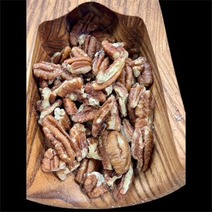 May include: A wooden bowl filled with pecan halves. The pecans are light brown with a slightly textured surface. The bowl is made of a light-colored wood with visible grain patterns. The background is black.