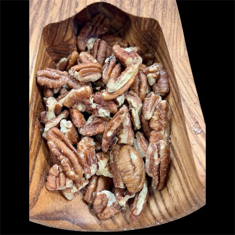 May include: A wooden bowl filled with pecan halves. The pecans are light brown with a slightly textured surface. The bowl is made of a light-colored wood with visible grain patterns. The background is black.