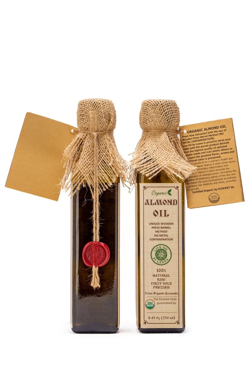 May include: Two glass bottles of organic almond oil with burlap coverings and red wax seals. The bottles are labelled with the words "Organic Almond Oil" and "Live Oil by Lesna".
