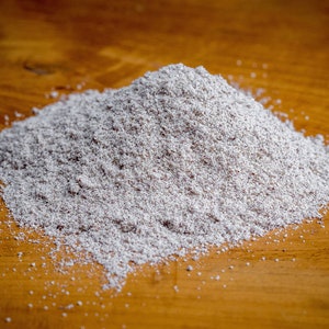 May include: A mound of fine, light gray, powdery substance with a grainy texture. The powder has a speckled appearance with tiny dark particles mixed throughout the light gray base, forming a conical pile with scattered grains at the base.