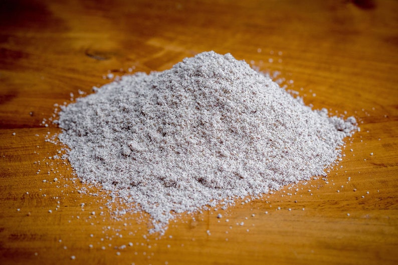 May include: A mound of fine, light gray, powdery substance with a grainy texture. The powder has a speckled appearance with tiny dark particles mixed throughout the light gray base, forming a conical pile with scattered grains at the base.