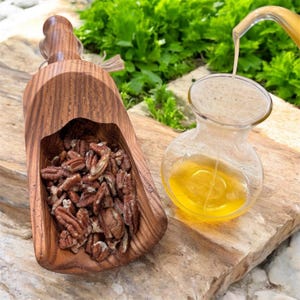 May include: A wooden scoop filled with pecans sits next to a small glass decanter being filled with oil. The oil is a golden yellow color. The background features green foliage.