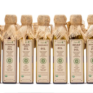 May include: A row of 12 glass bottles with burlap tops, each containing a different type of organic seed oil. The bottles are labeled with the type of oil, including almond, walnut, pumpkin, flax, hazelnut, hemp, mustard, poppy, and black sesame.