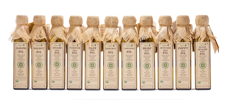 May include: A row of 12 glass bottles with burlap tops, each containing a different type of organic seed oil. The bottles are labeled with the type of oil, including almond, walnut, pumpkin, flax, hazelnut, hemp, mustard, poppy, and black sesame.