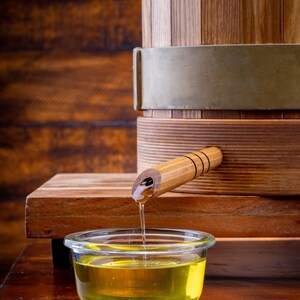 May include: A wooden barrel with metal bands and a spout dispensing a golden liquid into a clear glass bowl. The barrel sits on a wooden surface, with a wooden background.