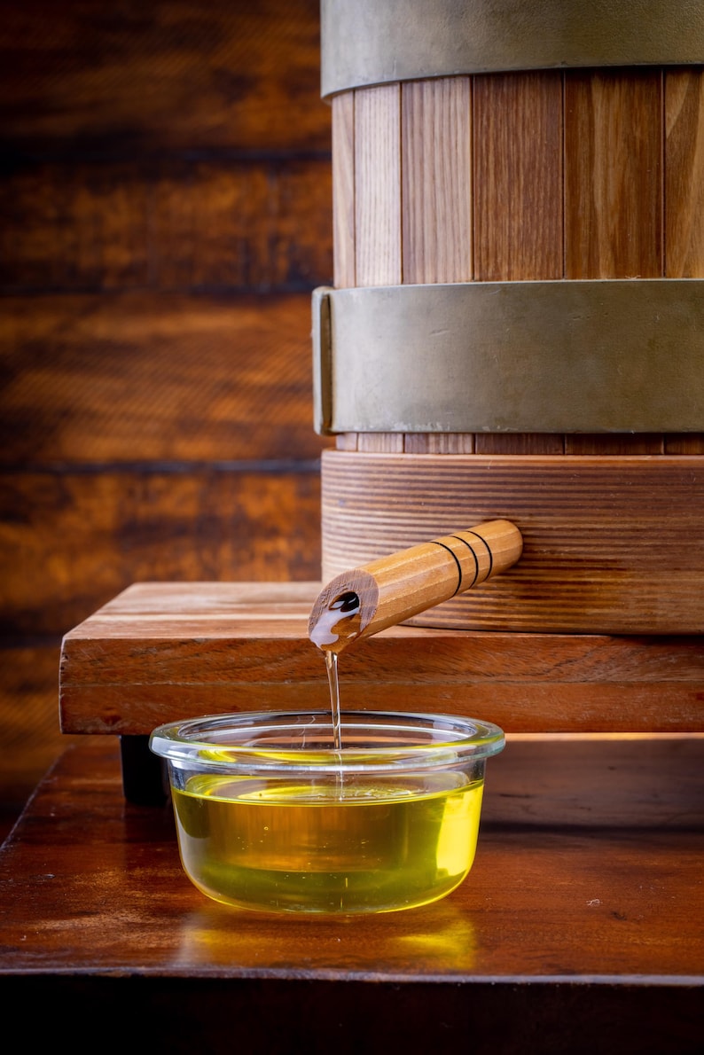 May include: A wooden barrel with metal bands and a spout dispensing a golden liquid into a clear glass bowl. The barrel sits on a wooden surface, with a wooden background.