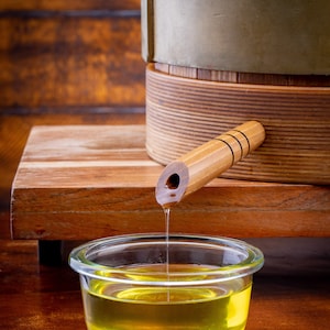 May include: A wooden barrel with a spout pouring golden yellow oil into a clear glass bowl. The barrel has a metal band around it and a wooden handle.