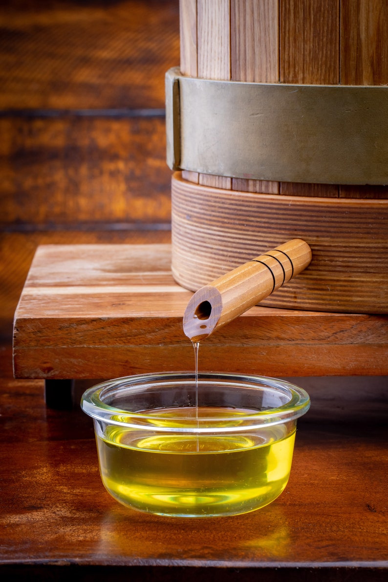 May include: A wooden barrel with a spout pouring golden yellow oil into a clear glass bowl. The barrel has a metal band around it and a wooden handle.