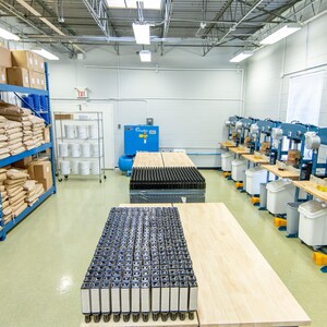 May include: An industrial warehouse interior with blue shelving filled with boxes and bags. Several machines are lined up along the wall, with work tables and containers. The floor is light green, and the ceiling has bright overhead lights.