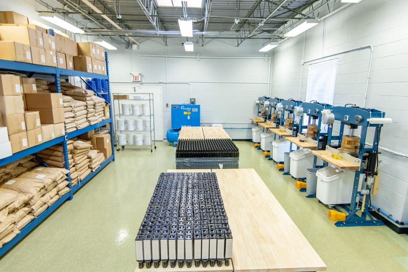 May include: An industrial warehouse interior with blue shelving filled with boxes and bags. Several machines are lined up along the wall, with work tables and containers. The floor is light green, and the ceiling has bright overhead lights.