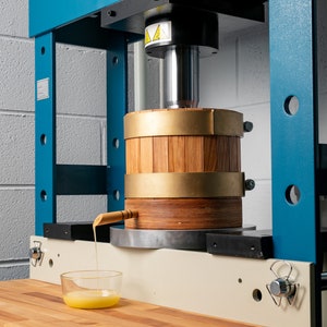 May include: A blue and beige industrial hydraulic press featuring a cylindrical wooden barrel with brass bands. A wooden spout extends from the barrel, dispensing a thick yellow liquid into a small, clear glass bowl positioned on a wooden surface.
