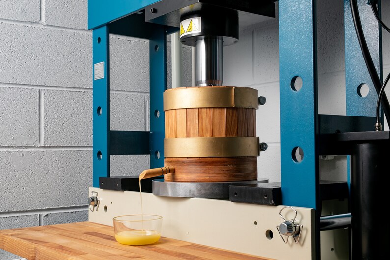 May include: A blue and beige industrial hydraulic press featuring a cylindrical wooden barrel with brass bands. A wooden spout extends from the barrel, dispensing a thick yellow liquid into a small, clear glass bowl positioned on a wooden surface.