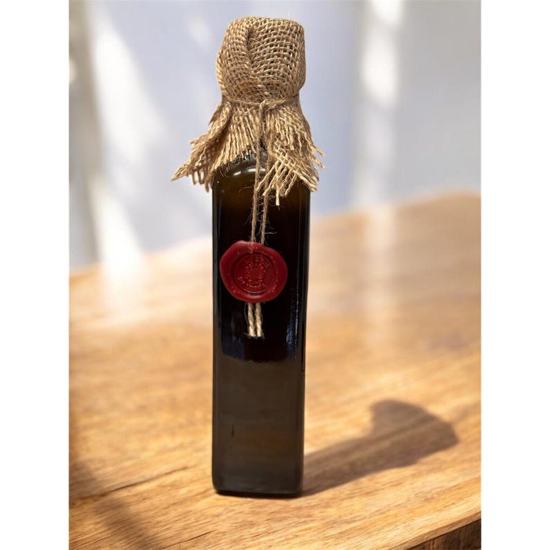 May include: A dark glass bottle with a burlap and twine closure, sealed with a red wax stamp. The bottle is rectangular and sits on a wooden surface. The overall aesthetic is rustic and artisanal.