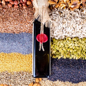 May include: A dark glass bottle with a burlap and twine closure, sealed with a red wax stamp, is surrounded by various seeds and nuts. The arrangement includes hazelnuts, flax seeds, poppy seeds, sunflower seeds, pumpkin seeds, and almonds.