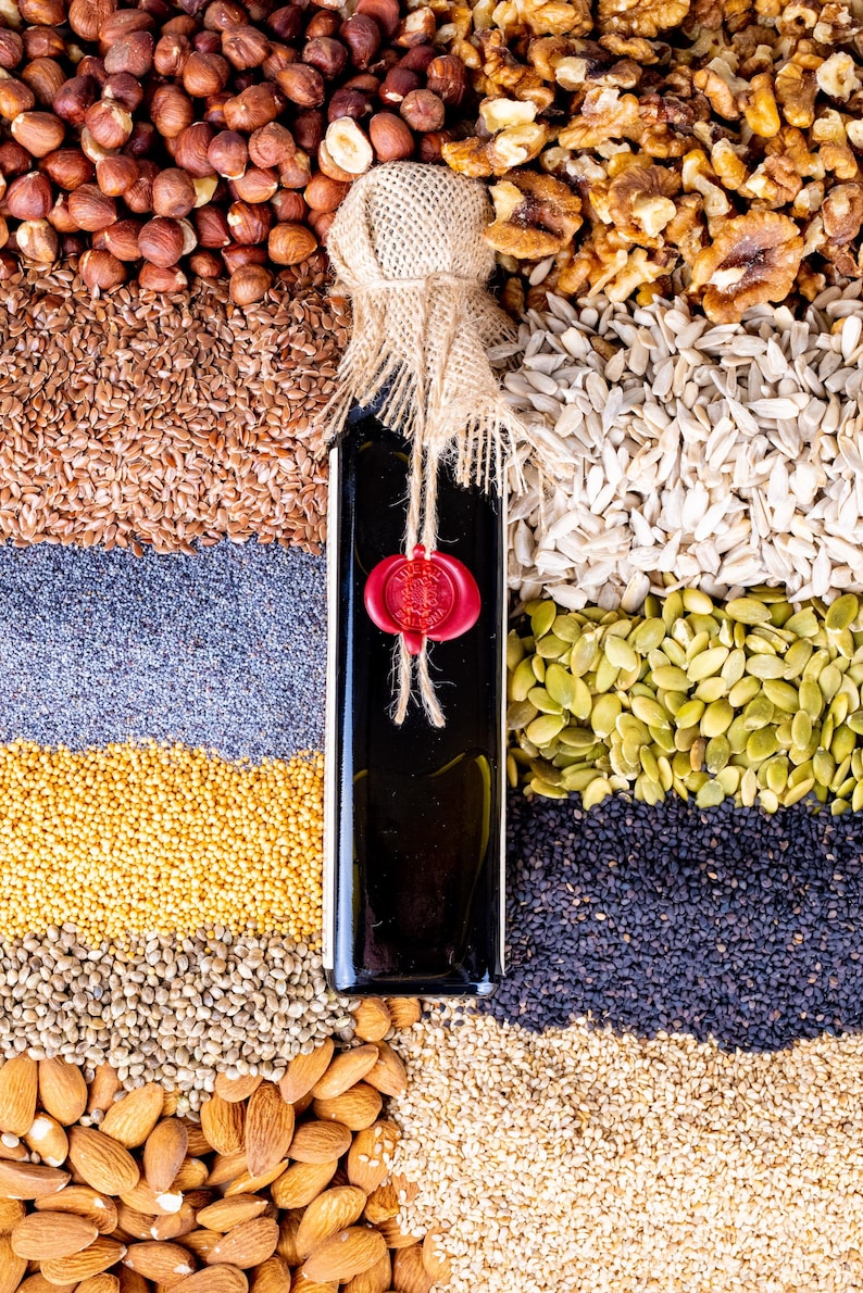 May include: A dark glass bottle with a burlap and twine closure, sealed with a red wax stamp, is surrounded by various seeds and nuts. The arrangement includes hazelnuts, flax seeds, poppy seeds, sunflower seeds, pumpkin seeds, and almonds.