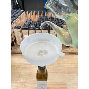 May include: A clear liquid is poured through a white funnel into a brown glass bottle labeled "Macadamia Oil." The funnel sits atop the bottle, filtering the liquid. Other bottles of oil are visible in the background.