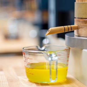 May include: A clear glass measuring cup with yellow liquid is positioned below a wooden spout. The spout is part of a wooden and metal apparatus. The liquid is dripping from the spout into the measuring cup. The cup is on a wooden surface.
