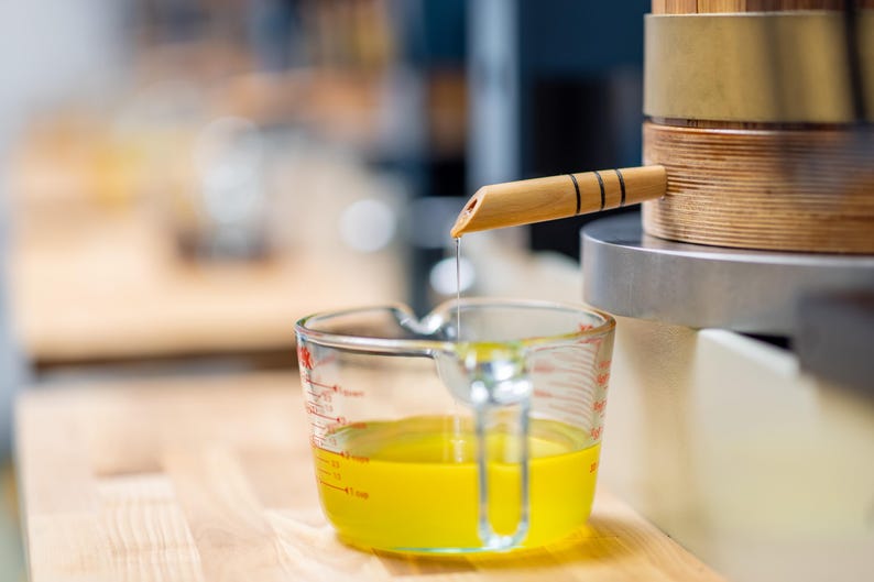 May include: A clear glass measuring cup with yellow liquid is positioned below a wooden spout. The spout is part of a wooden and metal apparatus. The liquid is dripping from the spout into the measuring cup. The cup is on a wooden surface.