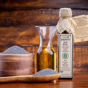 May include: A glass bottle of organic poppy seed oil with a burlap covering and a label that reads "Organic Poppy Seed Oil". The bottle is next to a wooden bowl filled with poppy seeds and a wooden spoon with more poppy seeds.