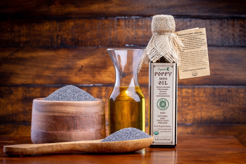 May include: A glass bottle of organic poppy seed oil with a burlap covering and a label that reads "Organic Poppy Seed Oil". The bottle is next to a wooden bowl filled with poppy seeds and a wooden spoon with more poppy seeds.