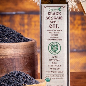 May include: A glass bottle of organic black sesame seed oil with a brown label that reads "Organic Black Sesame Seed Oil". The bottle is sitting on a wooden surface with a bowl of black sesame seeds in front of it.