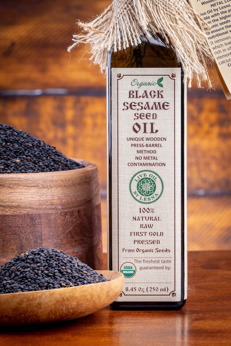 May include: A glass bottle of organic black sesame seed oil with a brown label that reads "Organic Black Sesame Seed Oil". The bottle is sitting on a wooden surface with a bowl of black sesame seeds in front of it.