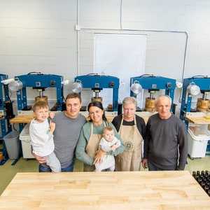 Peut inclure: Une famille de cinq personnes se tient devant une rang&eacute;e de presses industrielles bleues utilis&eacute;es pour la fabrication de cidre de pomme. La famille sourit et regarde la cam&eacute;ra. Il y a de nombreuses bouteilles en verre vides au premier plan.