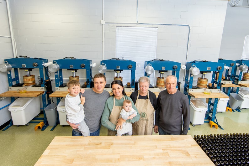 Peut inclure: Une famille de cinq personnes se tient devant une rang&eacute;e de presses industrielles bleues utilis&eacute;es pour la fabrication de cidre de pomme. La famille sourit et regarde la cam&eacute;ra. Il y a de nombreuses bouteilles en verre vides au premier plan.