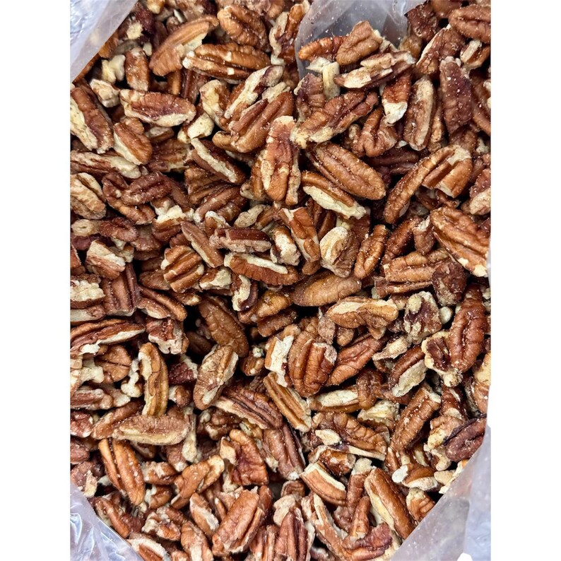 May include: A close-up shot of a clear plastic bag filled with pecan halves. The pecans are a mix of light and dark brown, with a slightly textured surface. The nuts are arranged in a dense, overlapping pattern, showcasing their natural shape and color.