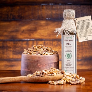 May include: A glass bottle of organic walnut oil with a burlap covering and a label that reads "Organic Walnut Oil". The bottle is next to a wooden bowl filled with walnut halves and a wooden spoon with more walnut halves.