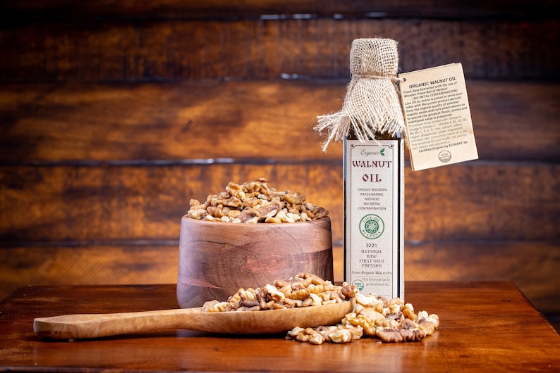 May include: A glass bottle of organic walnut oil with a burlap covering and a label that reads "Organic Walnut Oil". The bottle is next to a wooden bowl filled with walnut halves and a wooden spoon with more walnut halves.