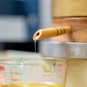 May include: A light-colored wooden spigot with three dark horizontal bands protruding from a wooden barrel. A thin, continuous stream of golden liquid flows from the spigot into a clear glass measuring cup with red markings below.