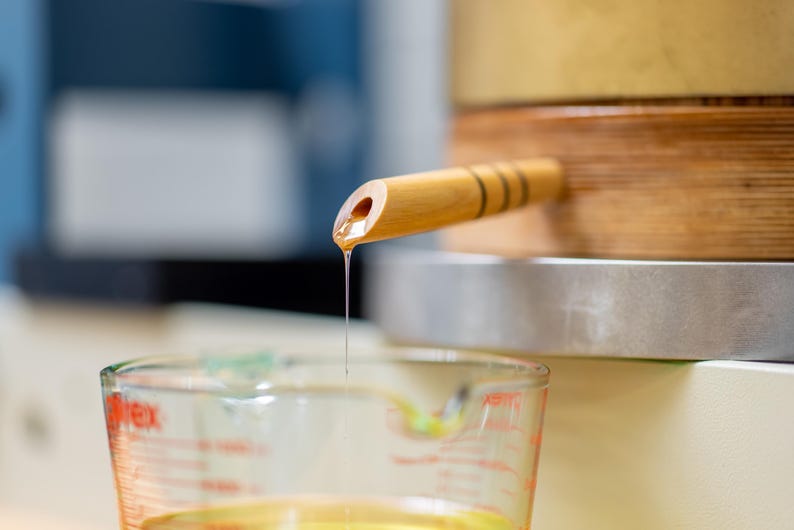 May include: A light-colored wooden spigot with three dark horizontal bands protruding from a wooden barrel. A thin, continuous stream of golden liquid flows from the spigot into a clear glass measuring cup with red markings below.