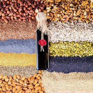May include: A rectangular glass bottle filled with dark liquid, topped with a burlap-wrapped cork and a red wax seal. The bottle is positioned vertically amidst horizontal layers of various nuts, seeds, and grains including hazelnuts, walnuts, and almonds.