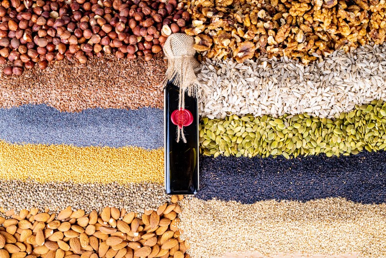 May include: A rectangular glass bottle filled with dark liquid, topped with a burlap-wrapped cork and a red wax seal. The bottle is positioned vertically amidst horizontal layers of various nuts, seeds, and grains including hazelnuts, walnuts, and almonds.