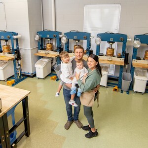 May include: A family of four stands in a workshop with multiple industrial-style presses. The parents are holding their two young children. The workshop has a light green tiled floor and a wooden table with many small black boxes.