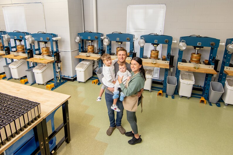 May include: A family of four stands in a workshop with multiple industrial-style presses. The parents are holding their two young children. The workshop has a light green tiled floor and a wooden table with many small black boxes.