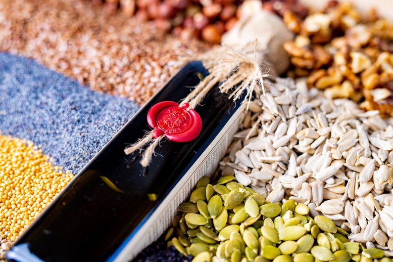 Peut inclure: Une bouteille en verre fonc&eacute; avec un sceau de cire rouge et de la ficelle attach&eacute;e autour du cou. La bouteille est entour&eacute;e de diverses graines et noix, notamment des graines de tournesol, des graines de citrouille, des graines de pavot, des graines de lin et des noix.