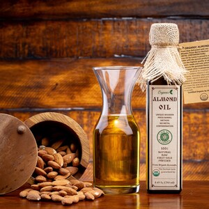 May include: A glass bottle of organic almond oil with a hessian lid. The bottle is labelled with the words "Organic Almond Oil" and "100% Natural Raw First Cold Pressed". The bottle is sitting on a wooden surface next to a wooden bowl filled with almonds.