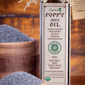 May include: A glass bottle of organic poppy seed oil with a brown label. The label reads "Organic Poppy Seed Oil", "Unique Wooden Press-Barrel Method", "No Metal Contamination", "100% Natural Raw First Gold Pressed", "From Organic Seeds", "The freshest taste guaranteed by:", and "8.45 oz (250 ml)". A wooden bowl filled with poppy seeds is in the foreground.
