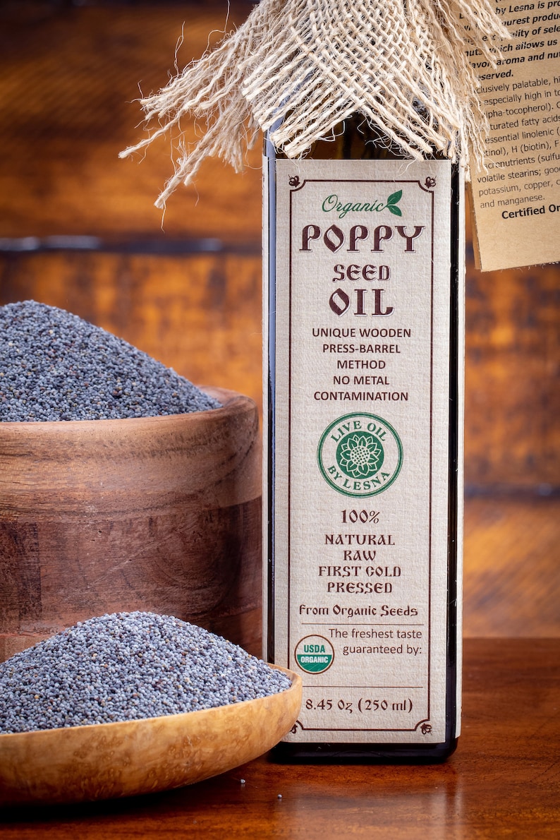 May include: A glass bottle of organic poppy seed oil with a brown label. The label reads "Organic Poppy Seed Oil", "Unique Wooden Press-Barrel Method", "No Metal Contamination", "100% Natural Raw First Gold Pressed", "From Organic Seeds", "The freshest taste guaranteed by:", and "8.45 oz (250 ml)". A wooden bowl filled with poppy seeds is in the foreground.