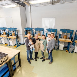 May include: A family of five stands in front of a row of blue hydraulic presses used for making oil. The presses are on a light green floor. The family is smiling and holding a baby. There are several white plastic bins in front of the presses.