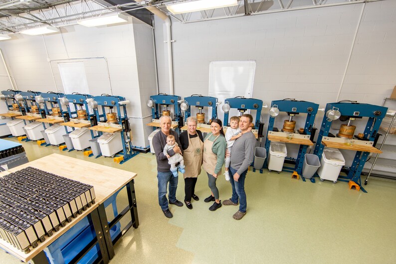 May include: A family of five stands in front of a row of blue hydraulic presses used for making oil. The presses are on a light green floor. The family is smiling and holding a baby. There are several white plastic bins in front of the presses.