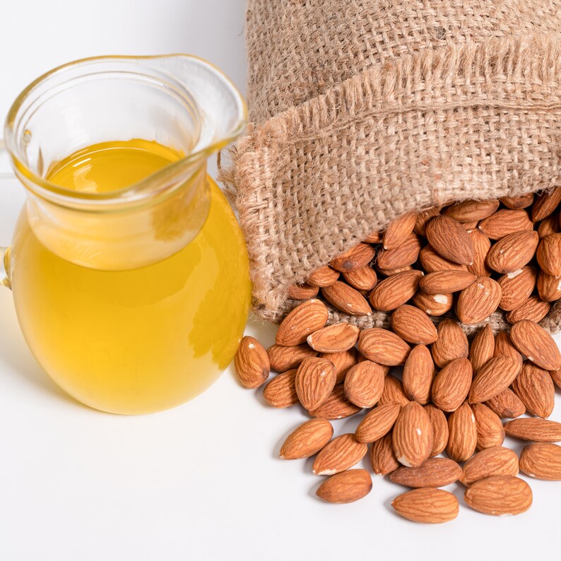 May include: A glass jug filled with light yellow almond oil next to a hessian sack spilling out almonds.