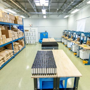May include: An industrial workshop with blue shelving filled with boxes and bags. Two wooden work tables are in the center, with a black grid of items on one. Several machines line the right wall.