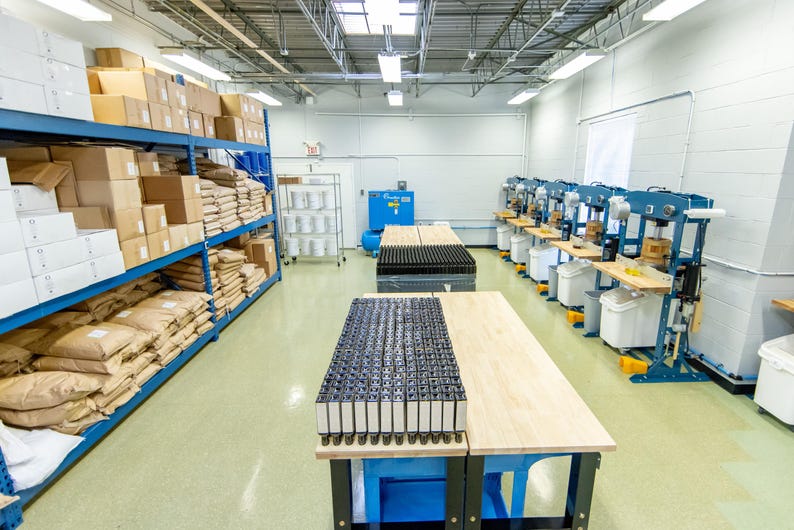 May include: An industrial workshop with blue shelving filled with boxes and bags. Two wooden work tables are in the center, with a black grid of items on one. Several machines line the right wall.
