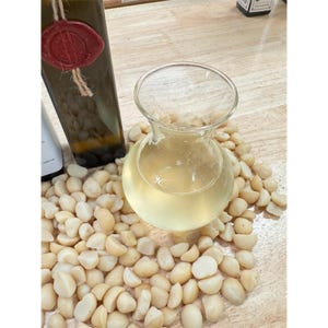 May include: A clear glass carafe filled with a light yellow liquid, surrounded by shelled macadamia nuts. A dark green bottle with a red wax seal is visible in the background. The scene is set on a light-colored wooden surface.